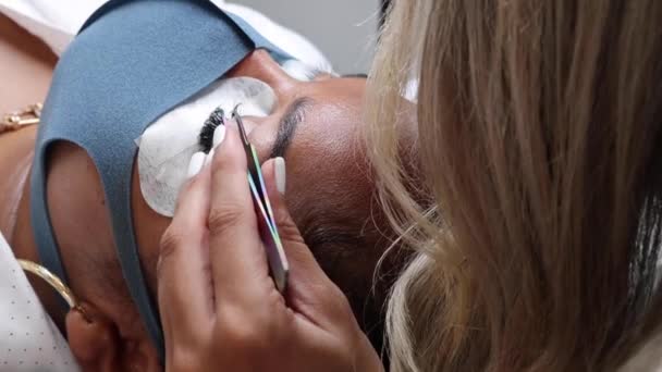cosmetologist que aplica pestanas falsas para a extensão no olho do cliente que usa a máscara protetora da cara no salão durante a pandemia covid 19 - Filmagem, Vídeo