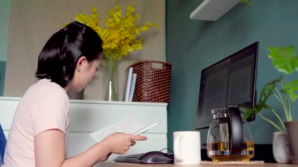 Πλευρική άποψη των ασιατικών γυναικών freelancer ανάγνωση σημειώσεις στο σημειωματάριο, ενώ κάθεται στο τραπέζι με τον υπολογιστή και εργάζονται για το έργο στο σπίτι - Πλάνα, βίντεο
