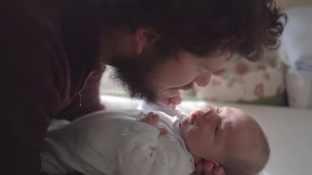 Zijaanzicht van volwassen bebaarde vader met klein kind leunend naar voren aan de zuigelingentafel terwijl ze naar elkaar kijken binnenshuis - Video