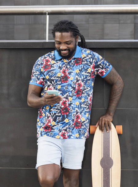 Alegre barbudo hombre afroamericano en ropa casual surfeando teléfono celular mientras está de pie cerca de la pared del edificio con longboard en la calle de la ciudad - Foto, imagen