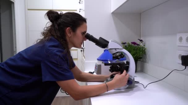 Vista lateral del veterinario femenino en uniforme azul mirando a través del microscopio mientras trabaja en laboratorio - Metraje, vídeo