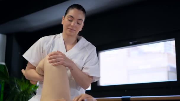 Happy masseuse in white robe massaging leg of anonymous female patient during physiotherapy session in clinic - Footage, Video