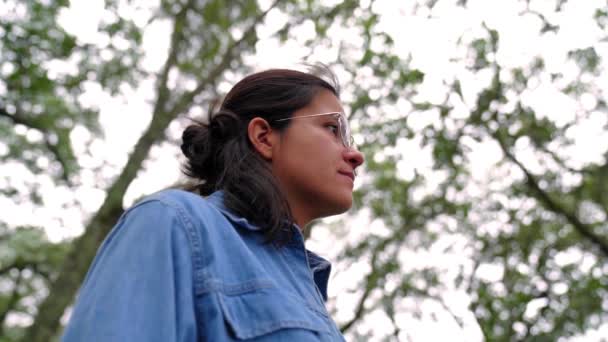 Zdola pozitivní etnická žena v brýlích s džínovou košilí stojící mezi stromy a odvracející se se zubatým úsměvem - Záběry, video
