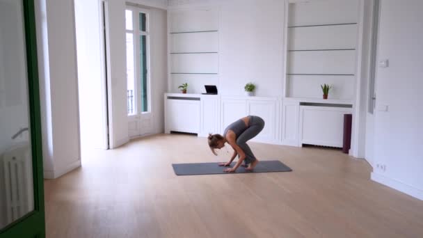 Гибкая женщина практикует йогу в позе журавля и стойке на голове, балансируя на коврике дома - Кадры, видео