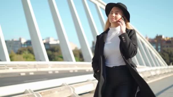 Młoda blondynka z plecakiem i kapeluszem rozmawiająca przez telefon podczas spaceru po asfaltowym moście w słoneczny dzień - Materiał filmowy, wideo