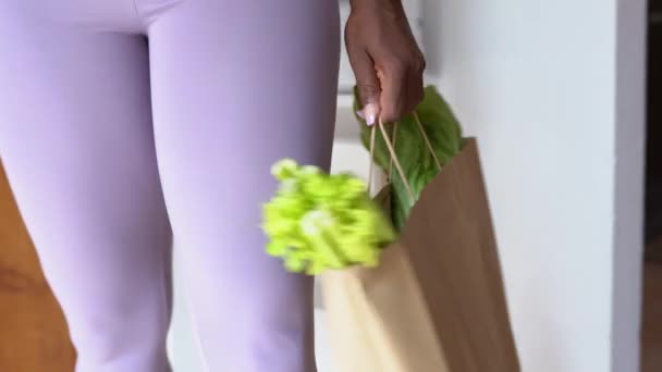 Zijaanzicht van gewas anonieme etnische vrouw met rijpe groene selderij en sla in boodschappentas staande in de keuken thuis - Video