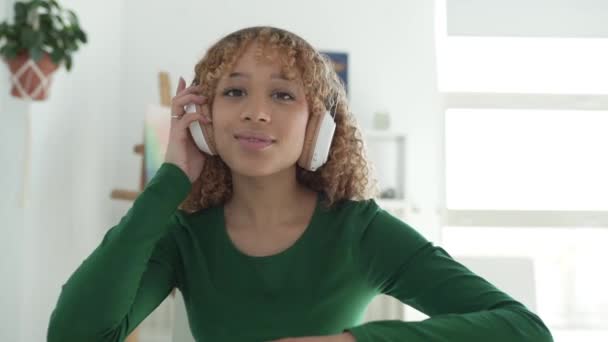 Joven mujer étnica con cabello rizado escuchando música de lista de reproducción en auriculares mientras está sentada en la mesa mientras hace vlog - Metraje, vídeo