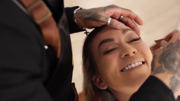 D'en haut sourire féminin heureux et montrant la langue pendant la session de maquillage avec visagiste tatoué dans le salon - Séquence, vidéo