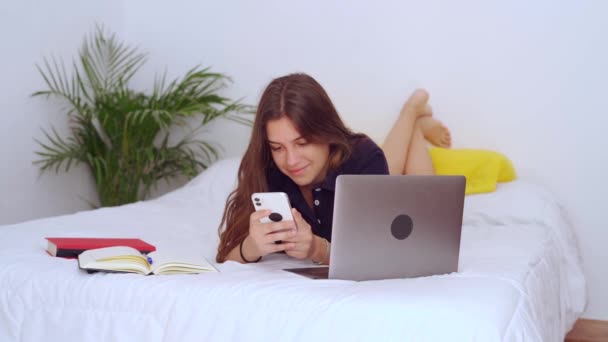 若い女性学生は、自宅でリモートオンライン研究中にノートパソコンや教科書やスマートフォン上のメッセージとベッドの上に横たわっています - 映像、動画