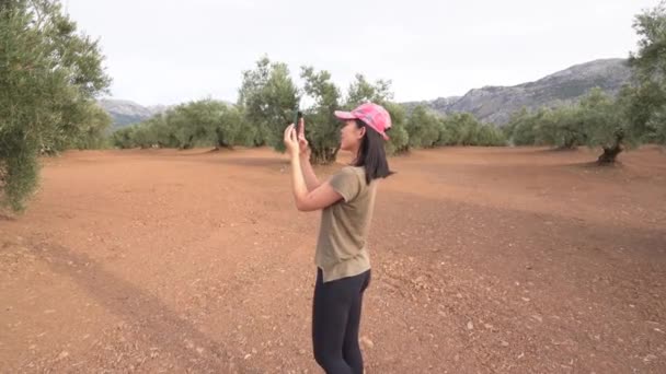 Nő sapkában rögzíti a videót a zöld olajfa a modern okostelefon, miközben bekapcsolja magát a ligetben - Felvétel, videó