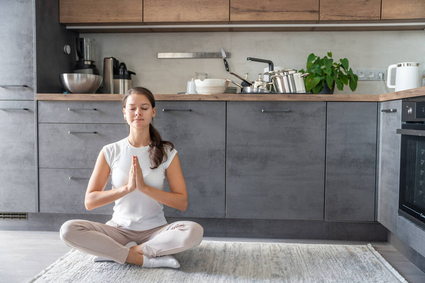 Young woman is sitting and doing meditation on background of dirty dishes in the kitchen. Slowdown, self-care, slow life concept. High quality photo - Fotografie, Obrázek