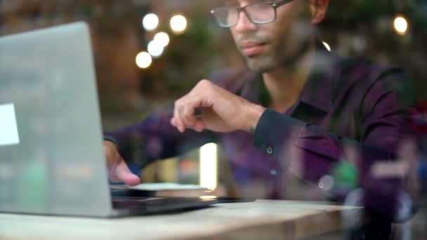 Poważny Latynoski menadżer w fioletowej koszuli i okularach, piszący na klawiaturze netbooka siedząc przy stole za oknem i pracując nad projektem w kawiarni - Materiał filmowy, wideo