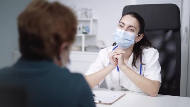 Jonge vrouw arts in wit uniform en stethoscoop op de nek te praten met oudere patiënt, bespreken gezondheidsproblemen schrijft klachten, vul medische formulier - Video