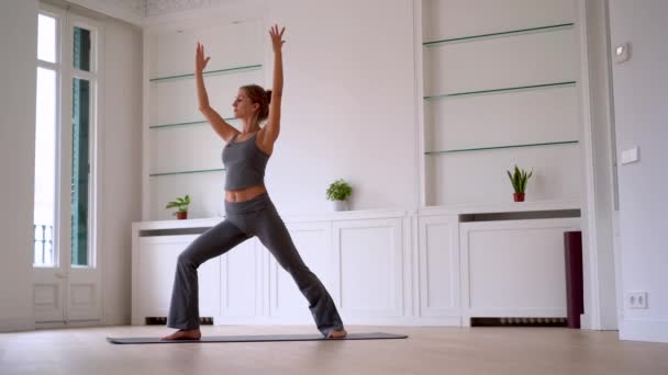 Vista laterale di femmina graziosa in abbigliamento sportivo praticare yoga mentre in piedi sul tappeto in camera spaziosa al mattino - Filmati, video