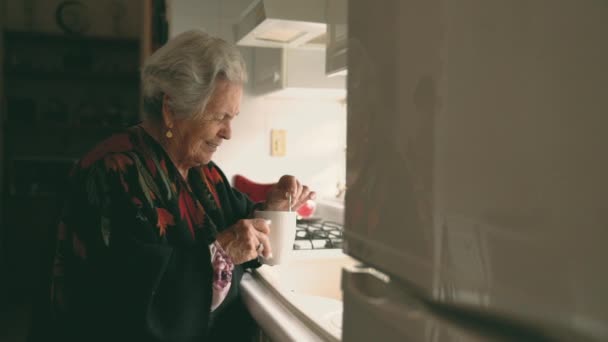 Vue latérale d'une femme âgée souriante dans un châle chaud avec une tasse debout près d'un évier blanc dans une cuisine blanche claire - Séquence, vidéo
