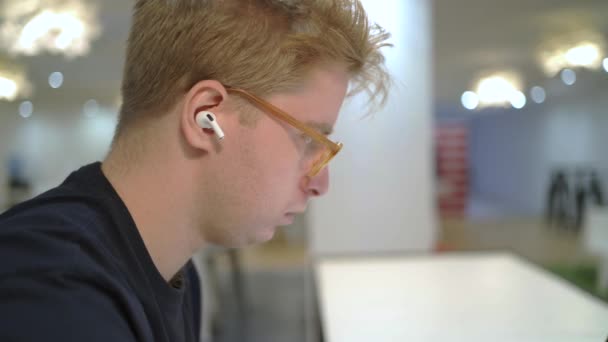 Πλευρική άποψη συμπυκνωμένου εργαζόμενου σε ασύρματα ακουστικά που εργάζεται σε φορητό υπολογιστή και χρησιμοποιεί κινητό τηλέφωνο σε μεσιτική εταιρεία - Πλάνα, βίντεο