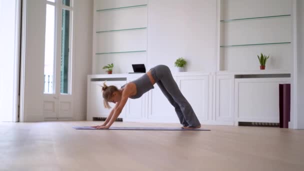 Widok z boku szczupła kobieta robi jogę na macie w przestronnym pokoju w domu - Materiał filmowy, wideo