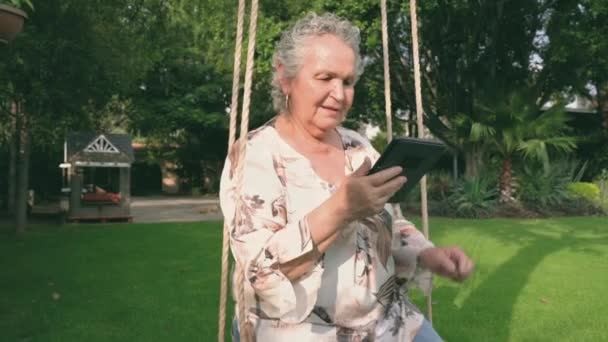 Старшая леди в блузке сидит на качелях в парке и читает электронную книгу - Кадры, видео