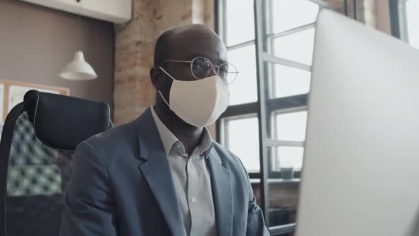 Alacsony szögű férfi afro-amerikai irodai munkás maszkot visel, nézi a képernyőn a számítógép és gépel valamit - Felvétel, videó