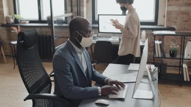 Maske takan ve bilgisayarda çalışan Afrika kökenli Amerikalı bir adamın orta eğim fotoğrafı. Maskeli genç beyaz bir kadın, elinde tabletle ofise gidiyor. - Video, Çekim