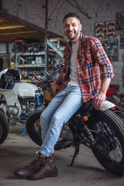 Bonito homem em roupas casuais está olhando para a câmera e sorrindo enquanto sentado na motocicleta na oficina
 - Foto, Imagem