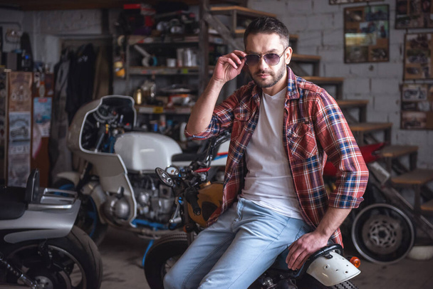 Przystojny stylowy mężczyzna okulary przeciwsłoneczne patrzy aparat siedząc na motocyklu w warsztacie - Zdjęcie, obraz