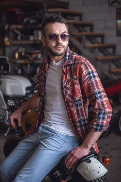 Bello uomo elegante in occhiali da sole è seduto sulla moto nel negozio di riparazione
 - Foto, immagini