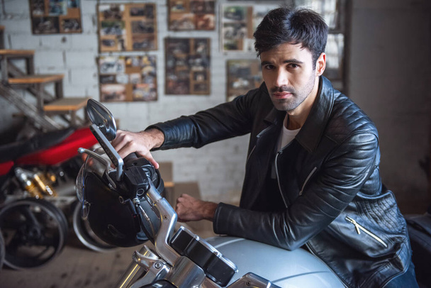 attraktiver Kerl in Lederjacke blickt in die Kamera, während er in der Werkstatt auf dem Motorrad sitzt - Foto, Bild
