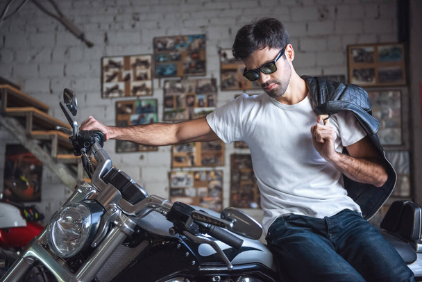 Atrakcyjny facet pewnie w okulary przeciwsłoneczne trzyma jego skórzaną kurtkę i patrząc na to siedząc na motocyklu w warsztacie - Zdjęcie, obraz
