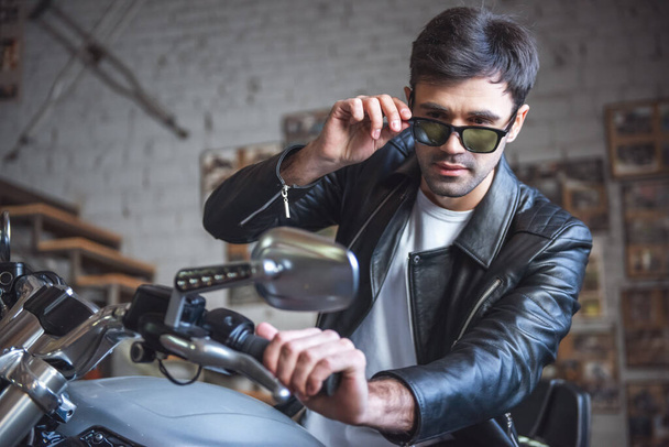 Attraente ragazzo elegante in giacca di pelle e occhiali da sole è seduto sulla moto in officina di riparazione
 - Foto, immagini