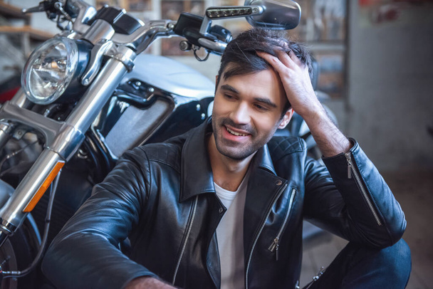 Atractivo tipo elegante en chaqueta de cuero está mirando hacia otro lado y sonriendo mientras está sentado cerca de la motocicleta en el taller de reparación
 - Foto, imagen