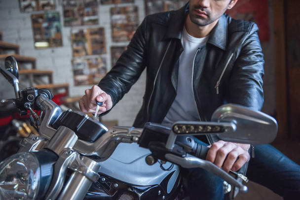 Atraente cara elegante em jaqueta de couro está segurando uma chave enquanto sentado na motocicleta na oficina
 - Foto, Imagem