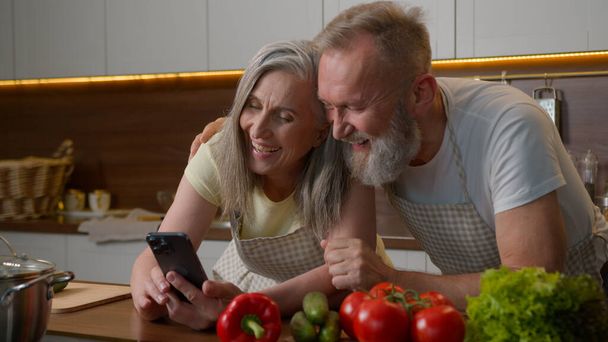 Nauraa eläkkeellä vanha harmaa tukka valkoihoinen perhe rakkaus pari matkapuhelimella verkossa tilata toimitus ruokaa vihanneksia keittiön pöydällä onnellinen vanhempi nainen ja mies puhuu ostoksia ruoanlaitto terveellistä ateriaa - Valokuva, kuva
