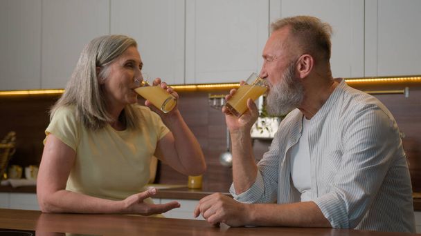 Happy middle-aged old senior couple Caucasian family woman and man drinking orange juice at kitchen home cheerful talk spouses retired wife and husband drink fresh fruit healthy nutrition healthcare - Photo, Image