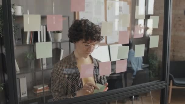 女性の白人マネージャーが昼間のオフィスに透明な壁に粘着性のあるノートを置く中間閉鎖 - 映像、動画