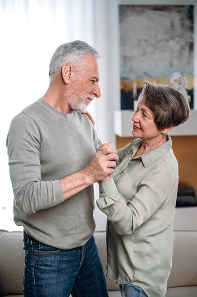 idős szerető pár táncol a nappaliban pihentető otthon hétvégén együtt. megértés, ragaszkodás, harmónia a házastársak között. függőleges lövés férfi és nő támogatja egymást nyugdíjas korban - Fotó, kép
