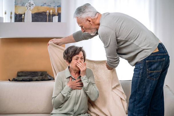 Amar hombre maduro de apoyo cuidar de su esposa enferma y anciana, cubriéndola con cuadros mientras ella está sentada en el sofá en casa, que sufre de la enfermedad por el virus de la gripe - Foto, imagen