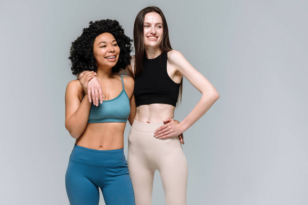 Ευρωπαϊκή κοπέλα με λεπτό σώμα αγκαλιά αφροαμερικάνικη σπορ γυναίκα μετά την προπόνηση στο γυμναστήριο. Δύο φίλες που στέκονται με στολή activewear απομονωμένες σε αντιγραφή γκρίζο φόντο στο γυμναστήριο μαζί - Φωτογραφία, εικόνα