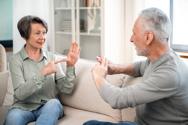 coppia anziana comunicare in lingua dei segni, mentre seduto sul divano a casa. anziana sordomuta e uomo che parla con le mani gesto e sorridente, capirsi - Foto, immagini