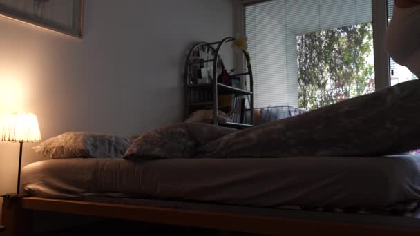 middle-aged woman, plump, plus size, in shorts, T-shirt, barefoot, with long blond hair, makes bed on bed in dark bedroom, with large window with blinds - Footage, Video