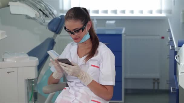 Atractiva dentista femenina usando teléfono celular en la clínica dental. Mujer bastante morena en uniforme médico blanco volteando en la pantalla táctil de su teléfono inteligente. Joven médico caucásico mirando copia
 - Metraje, vídeo