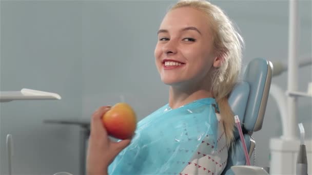Close up pretty blond woman okaying dentist checkup at the dental clinic. Attractive young woman showing her thumb up in dentist chair. Beautiful caucasian woman holding apple in her hand - Footage, Video