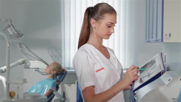 Kadın dişçinin dişçi dolabında bir şey işaretlediğini gösteren bir yan görüş. Tıbbi üniforma giymiş, elinde mavi kalemle beyaz bir kadın. Genç diş uzmanı. - Video, Çekim