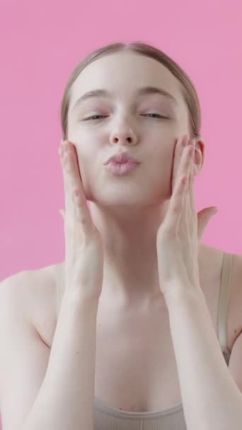 Όμορφη νεαρή κοπέλα με τέλειο δέρμα σε ροζ φόντο, χαμογελά θέτει και στέλνει φιλί αέρα. Έννοια της ευχαρίστησης αποτέλεσμα της χρήσης καλλυντικών προϊόντων φροντίδας του δέρματος Ροζ φόντο Συλλογή ομορφιάς Κάθετη βίντεο - Πλάνα, βίντεο