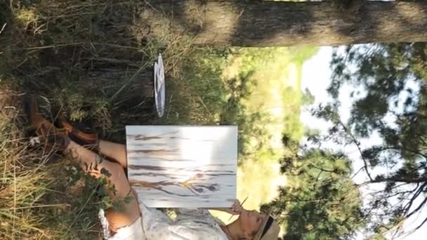 Μια καλλιτέχνης ζωγραφίζει μια εικόνα σε ένα πευκοδάσος. Όμορφη καλλιτέχνης κορίτσι σε εξωτερικούς χώρους. Ιστορικό τοπίο και δημιουργεί δημιουργικότητα στο δάσος το καλοκαίρι. Κάθετη βίντεο. - Πλάνα, βίντεο
