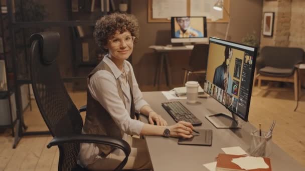 Dolly zoom retrato de la mujer caucásica gerente sonriendo, sosteniendo lápiz y sentado en el escritorio con el ordenador y cortes de imágenes en el monitor por la noche - Metraje, vídeo
