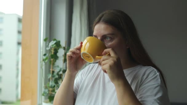 Piękna zrelaksowana kobieta z filiżanką herbaty lub kawy i patrząc przez okno siedząc na parapecie w domu. Wysokiej jakości materiał filmowy FullHD - Materiał filmowy, wideo