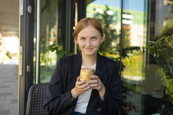 Крупним планом молода жінка в діловому повсякденному одязі п'є чашку кави, сидячи за невеликим столом і насолоджуючись часом у вуличному кафе. Високоякісна фотографія - Фото, зображення