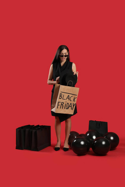 Шокированная молодая женщина с пакетами для покупок и воздушными шарами на красном фоне. Чёрная пятница - Фото, изображение