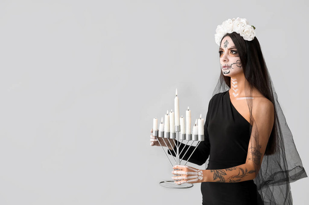 Mujer joven vestida de novia muerta para fiesta de Halloween sosteniendo candelabro con velas sobre fondo gris - Foto, imagen